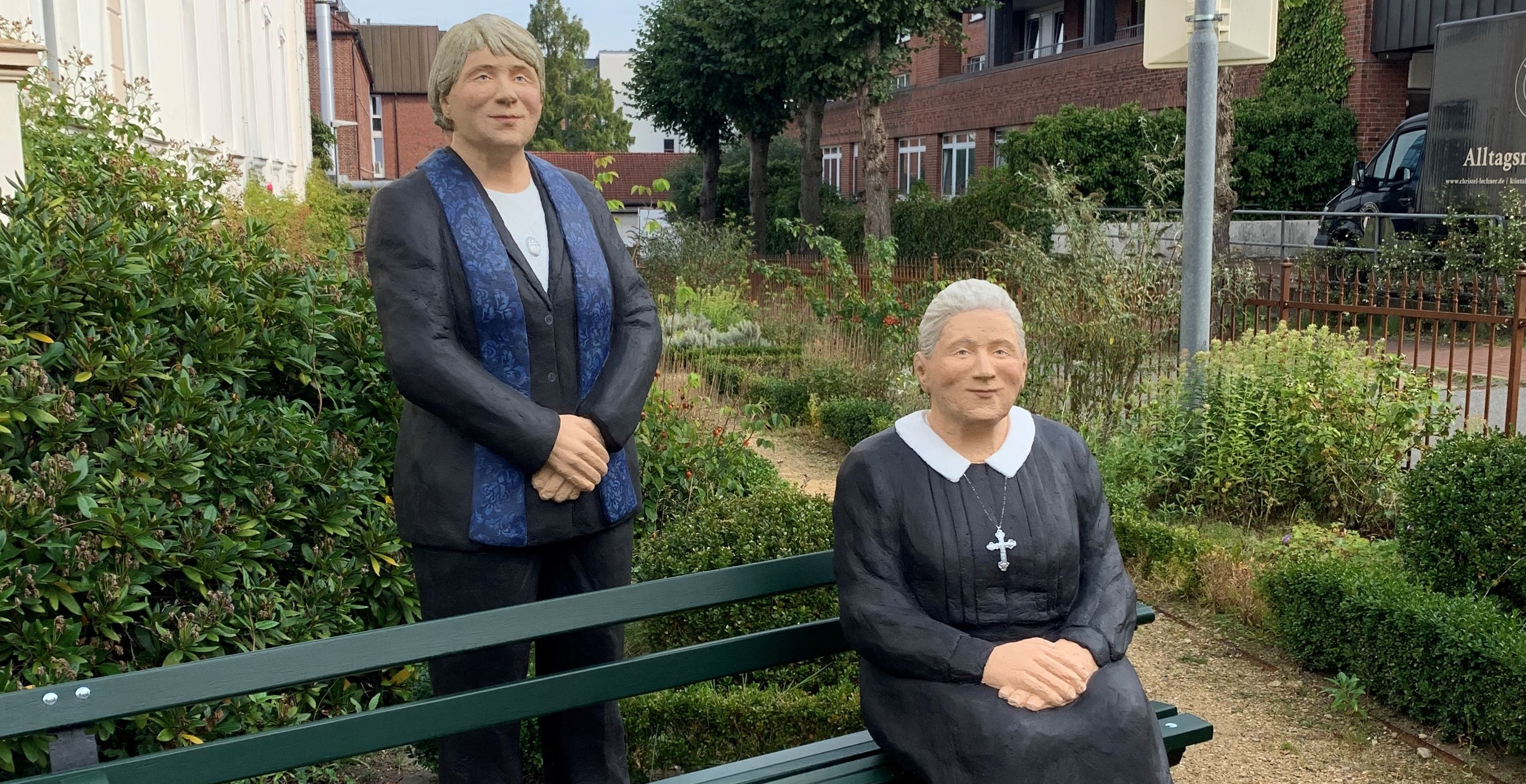 Zwei Skulpturen vor dem Haus Pniel: Sitzende Diakonisse mit Kreuz-Kette repräsentiert das Gestern, stehende Diakonisse Hosenanzug das Heute. Für das Morgen ist noch ein Platz auf oder hinter der Bank frei …